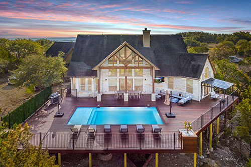 An aerial view of a vacation rental home showing a high-end home, a large wooden deck with a pool, and views of the surrounding Hill Country in Dripping Springs, Texas.