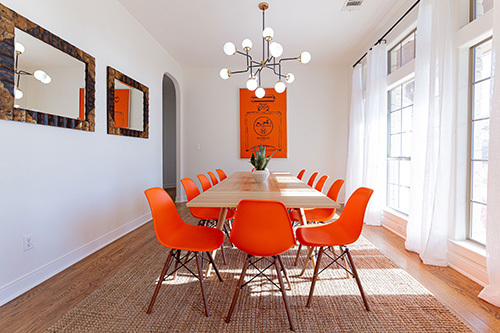 A light-filled dining room in a luxury vacation rental home showing a large table with orange chairs, a chandelier, and Hermès artwork.
