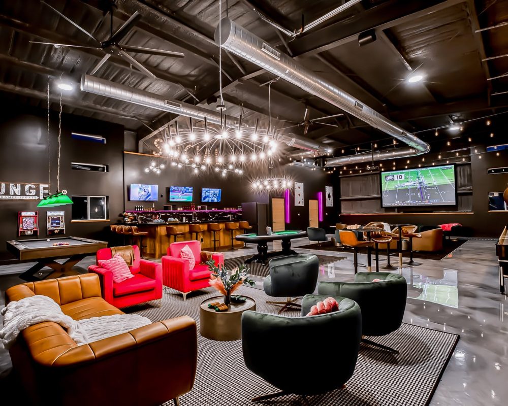 A luxurious vacation rental home hosted by GuestSpaces that features a large entertainment barn showing a projector screen, comfortable seating, a private bar, and a variety of game tables.